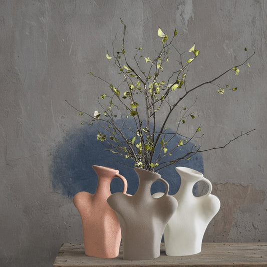 Three abstract vases in pink, white, and beige with a branch on a wooden surface against a gray wall.