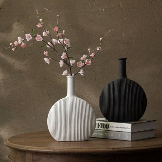 Two vases, one white with floral arrangement and the other black, on a wooden surface against a brown wall.