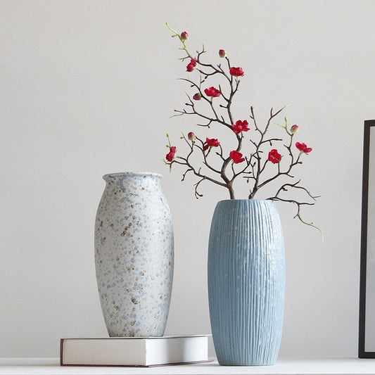 Two vases on a surface with a decorative branch with red flowers against a plain background.