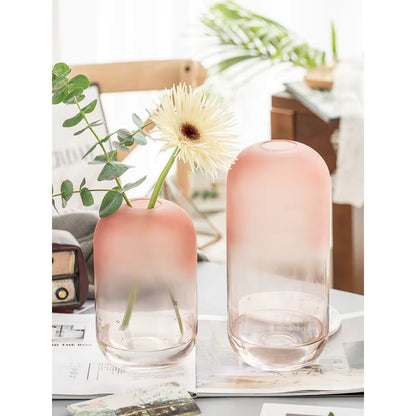 Two gradient pink vases with a single white flower on a table with a magazine and plant in the background.