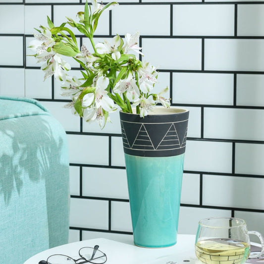 Turquoise vase with geometric design holding white flowers against a tiled wall.